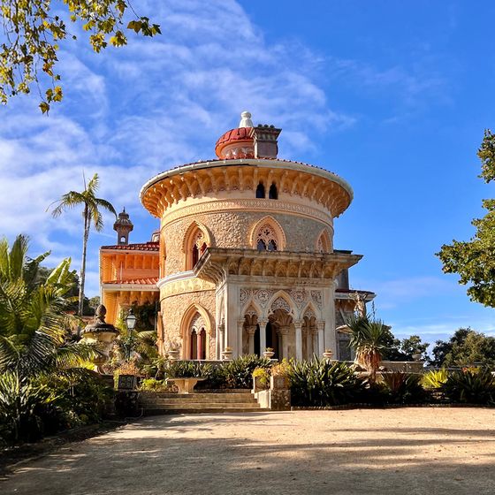 A historic palace with a round tower, ornate arches, and lush gardens under a blue sky.