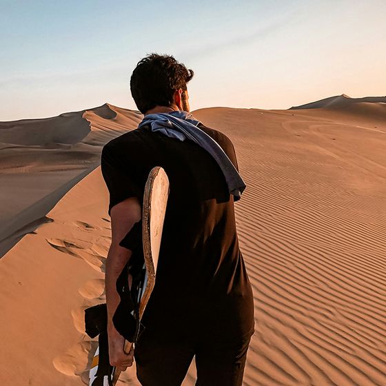 Man with skateboard walks on sand dunes at sunset, wearing a black shirt with a blue jacket draped over his shoulder.
