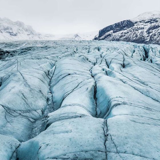 Close-up of a glacier's icy surface with visible cracks, extending toward snow-covered mountains under a cloudy sky.