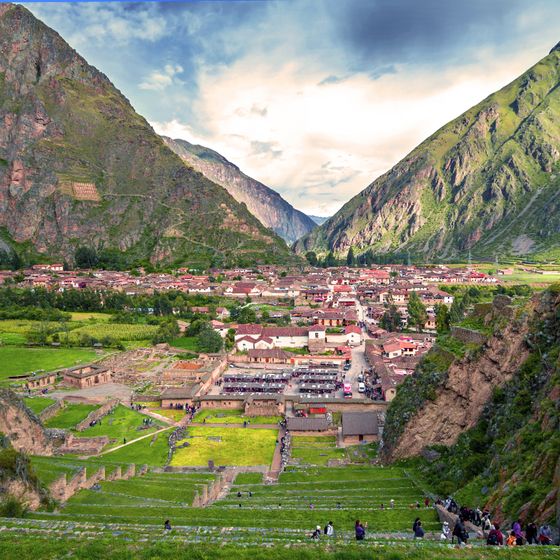 Scenic view of Ollantaytambo, Peru with ancient terraces, a valley, and distant mountains under a partly cloudy sky.