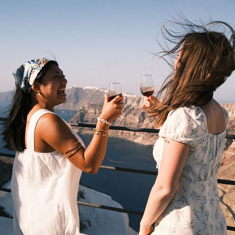 Two women clinking wine glasses on a sunny balcony overlooking the sea and mountains, with wind blowing their hair.