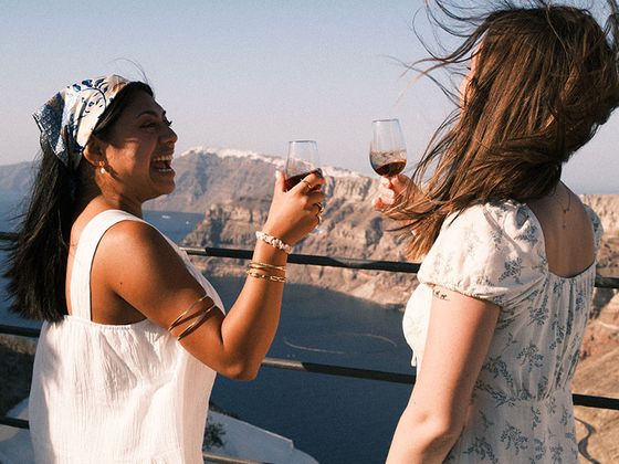 Two women enjoy glasses of wine with a scenic view of a coastal landscape and clear blue sky in the background.