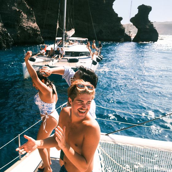 People enjoying a sunny day on a boat, dancing and smiling, with another boat and rocky cliffs in the background.