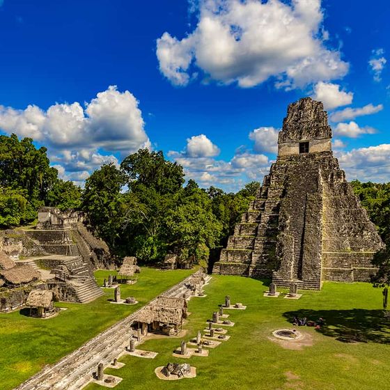 Ancient Mayan ruins with a stepped pyramid and stone structures surrounded by lush greenery under a blue sky with white clouds.