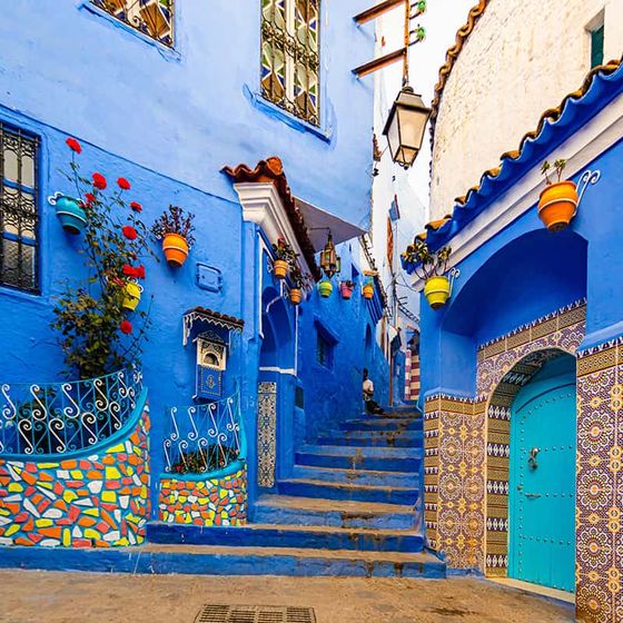 Vibrant blue Moroccan alley with colorful mosaic walls, a teal door, and potted plants hanging on walls, leading up stone steps.