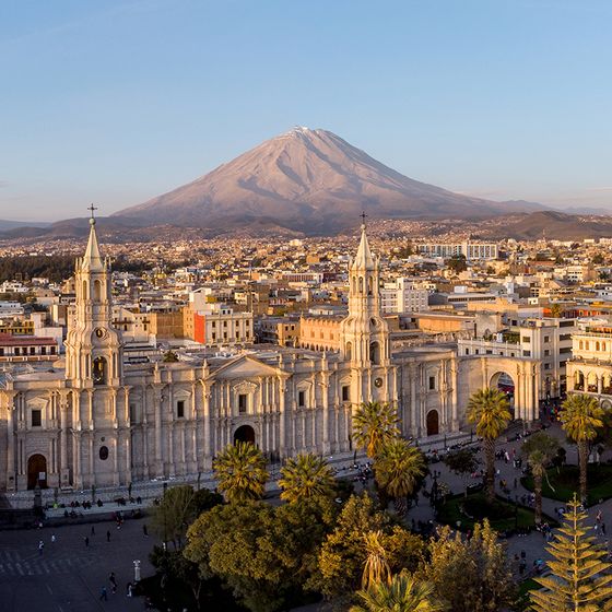 Aerial view of a grand white cathedral with two towers in a city, set against a backdrop of a snow-capped volcano and a clear blue sky.