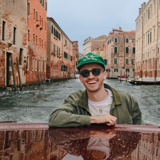Smiling person in sunglasses and green cap on a boat, with Venetian canal and historic buildings in the background.