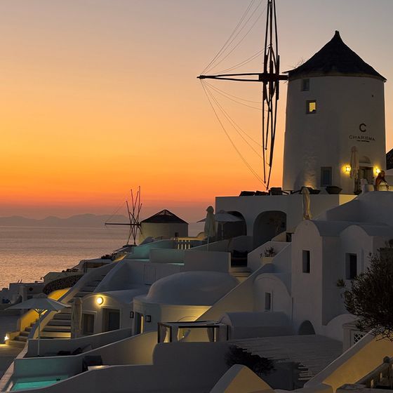 Sunset view of a traditional white windmill and buildings overlooking a calm sea in Santorini, Greece, with a warm, colorful sky.