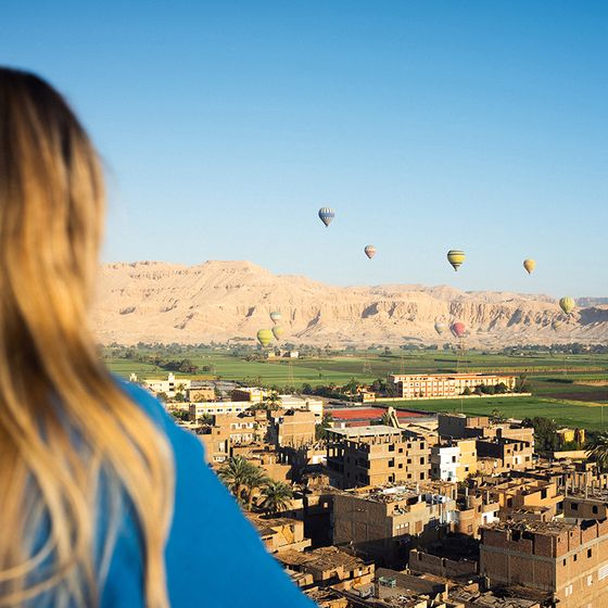 Person overlooks a town with hot air balloons in the sky, set against mountains and a clear blue sky.