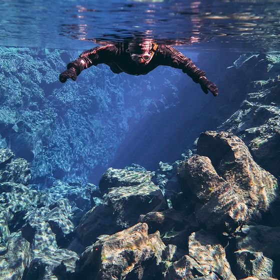 A diver in a wetsuit floats in clear blue water over rocky terrain, with sunlight streaming down, creating a serene underwater scene.