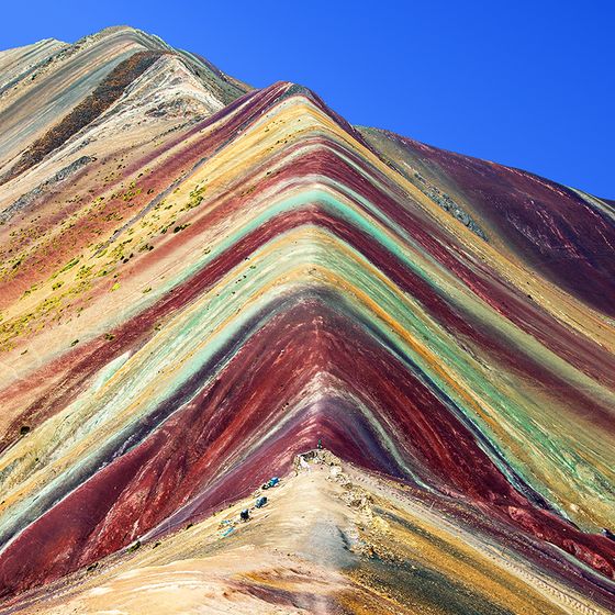 A vibrant, multicolored mountain peak with layers of red, yellow, and green against a clear blue sky.