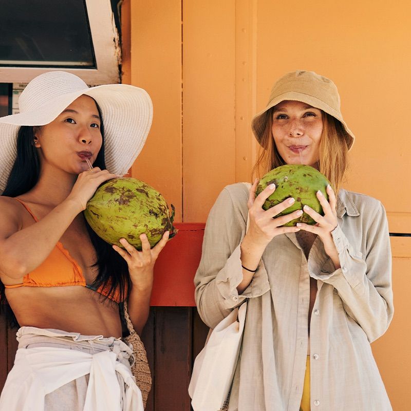 Two women in sun hats drink from coconuts, standing against an orange wall on a sunny day.