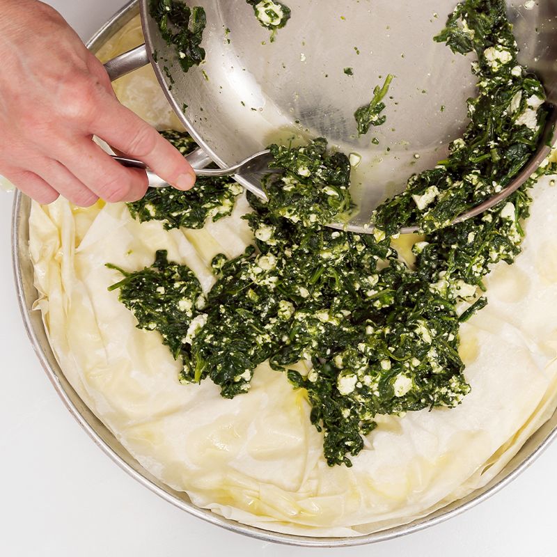 Person adding spinach and cheese mixture onto phyllo pastry in a baking pan.