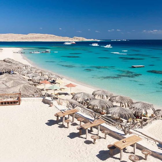 Tropical beach scene with straw umbrellas, wooden tables, clear turquoise water, and boats in the distance under a blue sky.