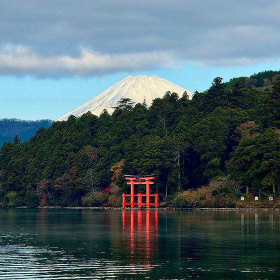 Red Torii gate on a calm lake, surrounded by dense trees, with a snow-capped Mount Fuji in the background under a cloudy sky.