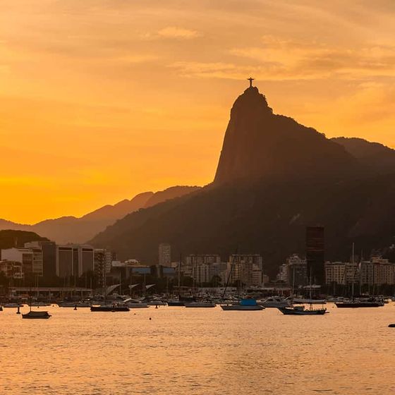 Sunset view over Rio de Janeiro's skyline with Sugarloaf Mountain silhouette and boats on the water.