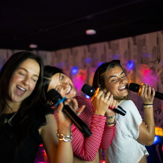 Three women joyfully singing into microphones together, with colorful lights and a patterned wall in the background.