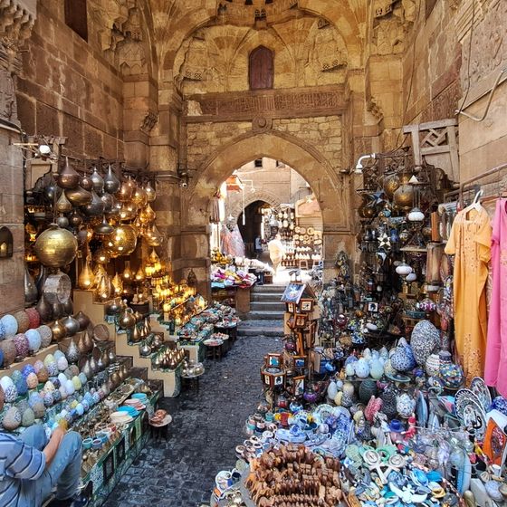 A bustling market alley with vibrant pottery, lanterns, and decorative items displayed on both sides beneath an arched stone entrance.