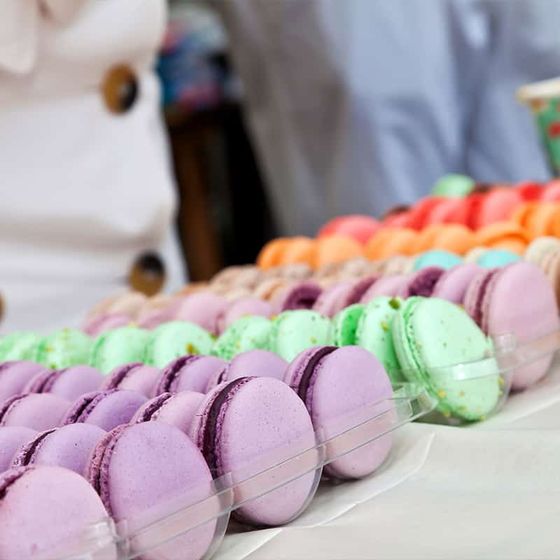 Colorful macarons arranged in rows on a clear tray, featuring shades of purple, green, orange, and pink. A person with a white shirt is nearby.