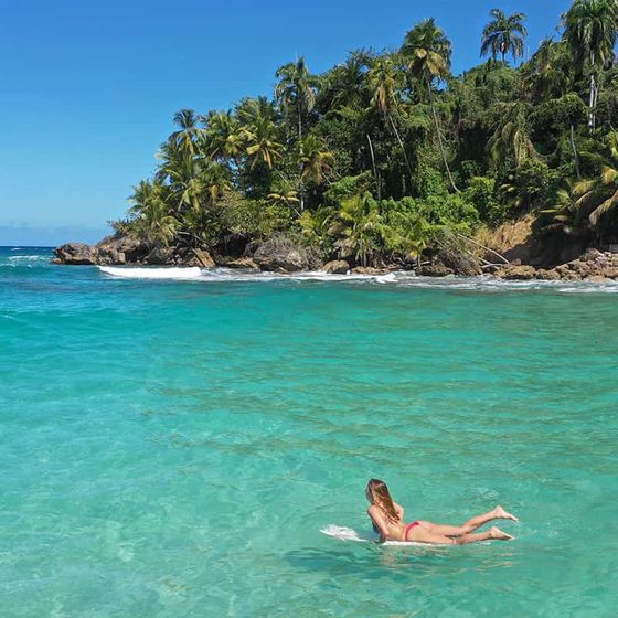 A person in a red swimsuit swims in clear turquoise water near a lush, palm-covered island under a clear blue sky.