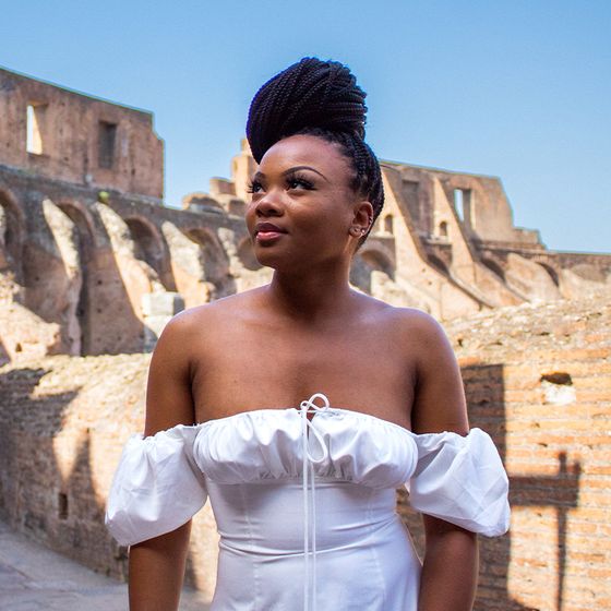 Woman in a white off-shoulder dress stands in front of ancient ruins under a clear blue sky.