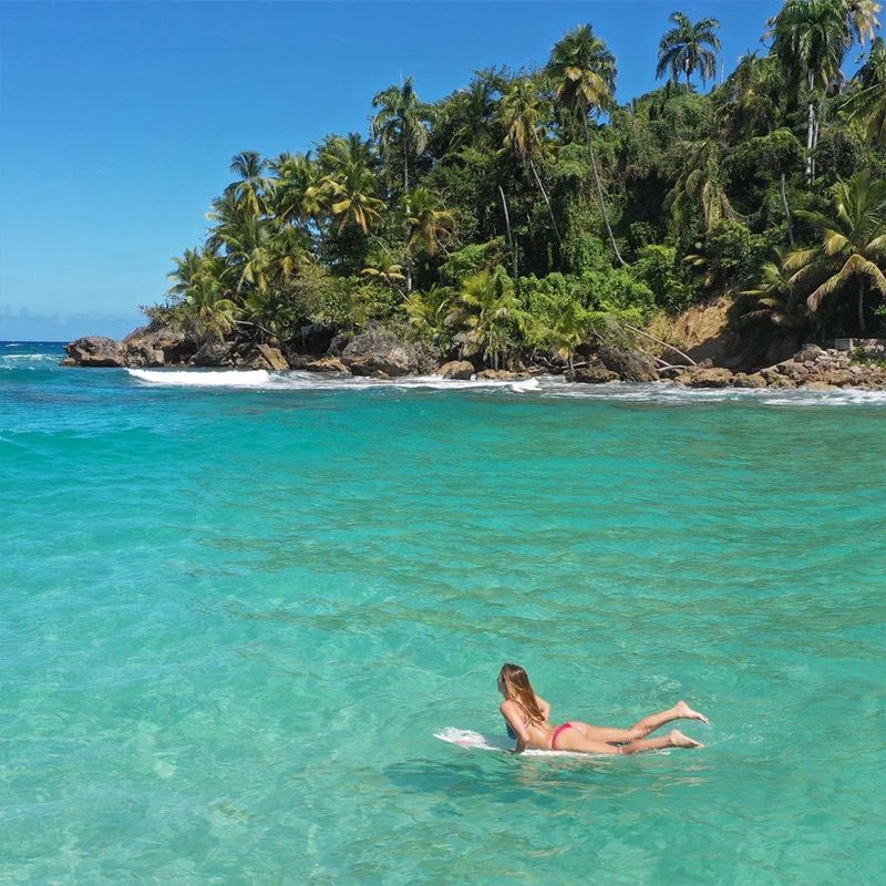 A person in a red swimsuit floats on clear turquoise water near a lush, tropical island with palm trees under a bright blue sky.