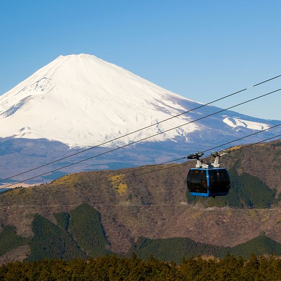 Cable car traversing above a forested landscape with a snow-capped mountain in the background under a clear blue sky.