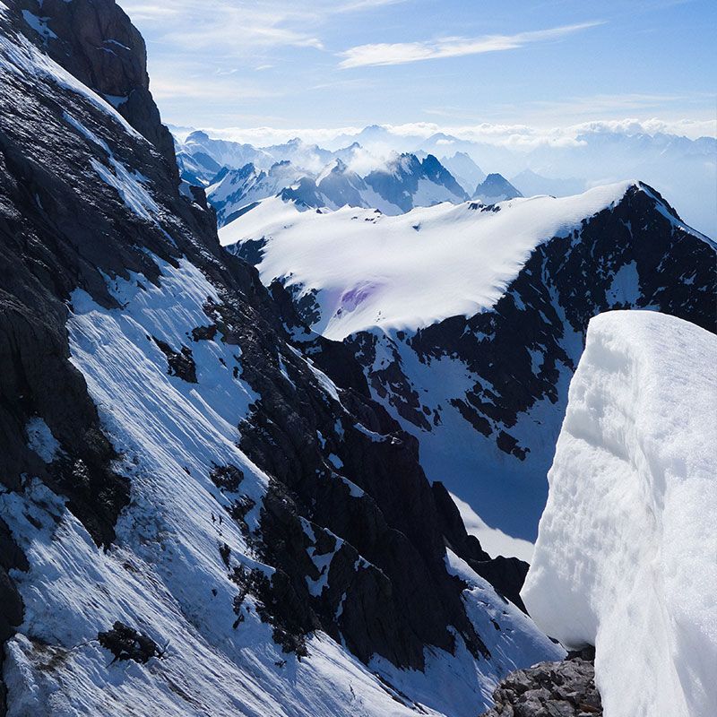 Snow-covered mountain peaks under a clear blue sky, with rugged cliffs and distant misty mountains in the background.