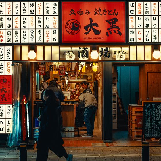 A brightly lit Japanese street food restaurant with people inside. The exterior is adorned with numerous signs featuring Japanese characters.
