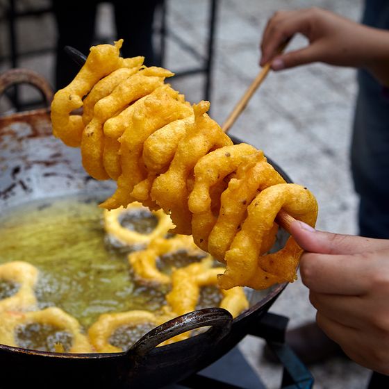 Close-up of hands frying crispy, golden fritters in hot oil. A skewer holds several freshly cooked fritters above a deep frying pan.