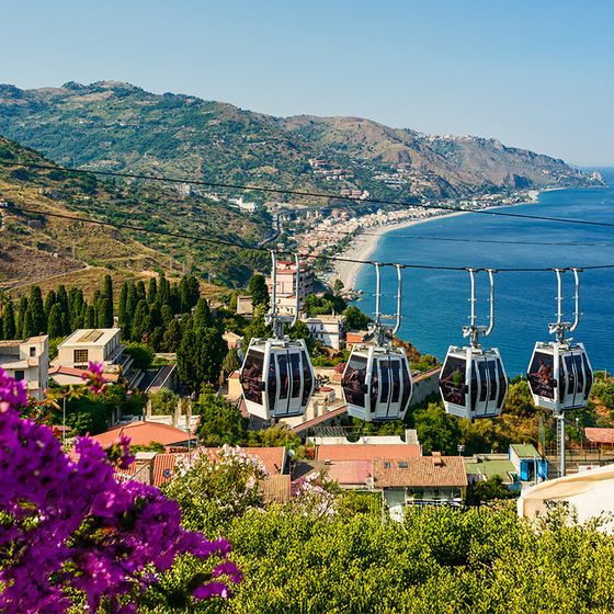 Cable cars over a scenic coastal landscape with blue sea and mountains, vibrant flowers in the foreground, and a distant town along the shoreline.