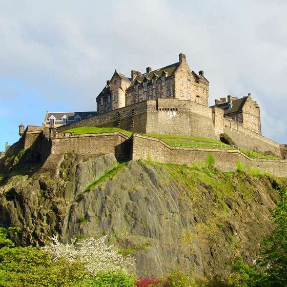 A historic stone castle sits atop a rocky hill, surrounded by lush greenery under a partly cloudy sky.