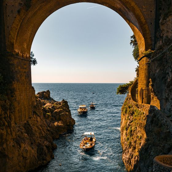 Boats navigating through a rocky cove under a large stone arch, with a calm sea and clear blue sky in the background.