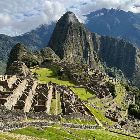 Ancient ruins of Machu Picchu with terraced fields and steep, lush mountain backdrop under a partly cloudy sky.