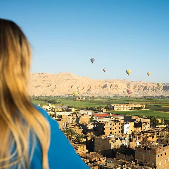 A person with long hair overlooks a landscape with multiple hot air balloons floating over a village and green fields, set against desert mountains.