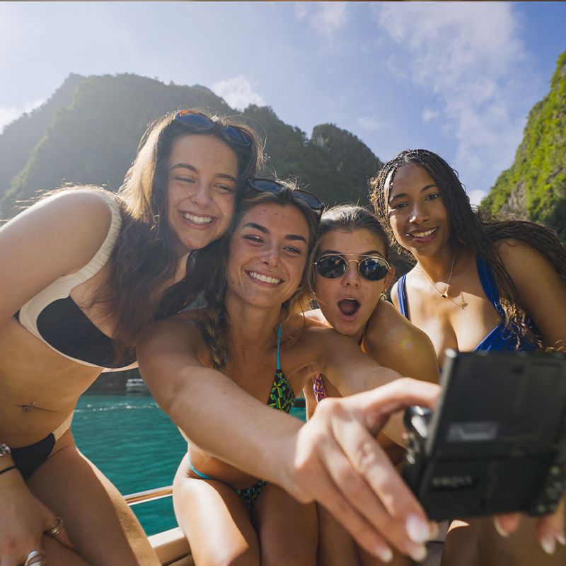 Four friends in swimwear take a selfie on a boat with lush green cliffs and clear blue water in the background.