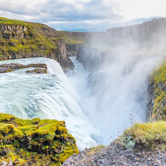 Powerful waterfall cascading through a rocky gorge with lush green cliffs under a cloudy sky.