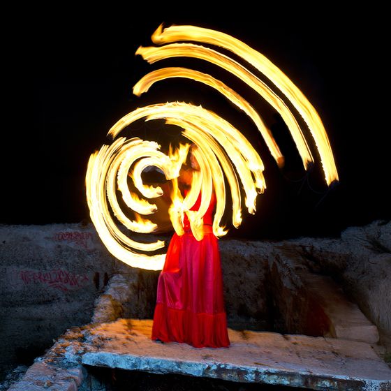 Person in a red dress spinning fiery light trails in spirals against a dark background, creating a dynamic, glowing effect.