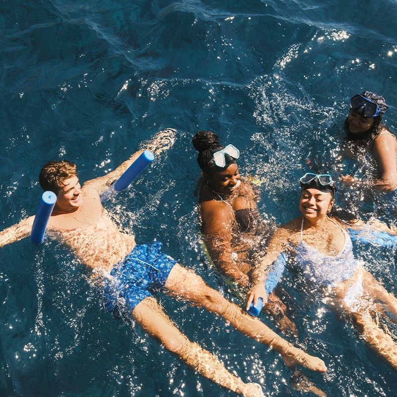 Four people in swimwear and goggles enjoying swimming in clear blue water, with two using pool noodles for support.