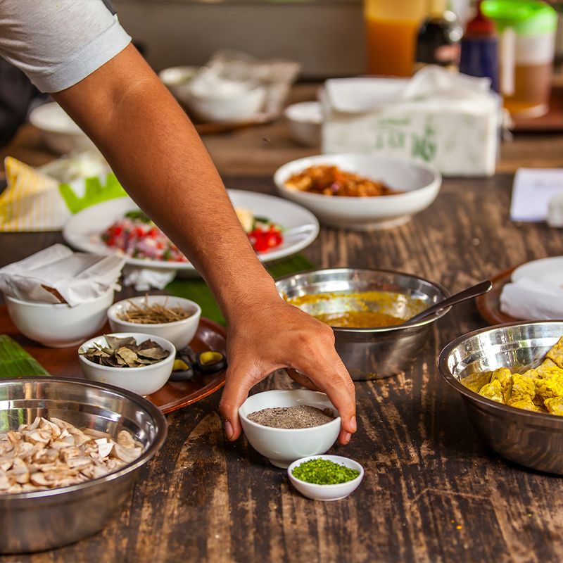 A person arranges small bowls of spices and ingredients on a wooden table, preparing a meal. Various dishes and condiments are in the background.