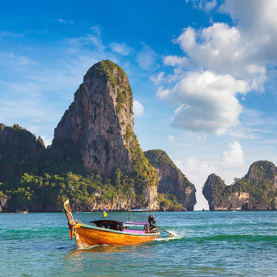 A traditional long-tail boat sails on turquoise waters with lush limestone cliffs and a clear blue sky in the background.
