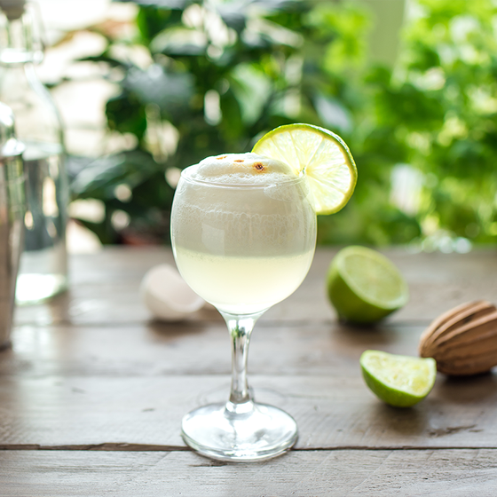 A frothy cocktail with a lime slice garnish in a wine glass on a wooden table, surrounded by limes and greenery in the background.