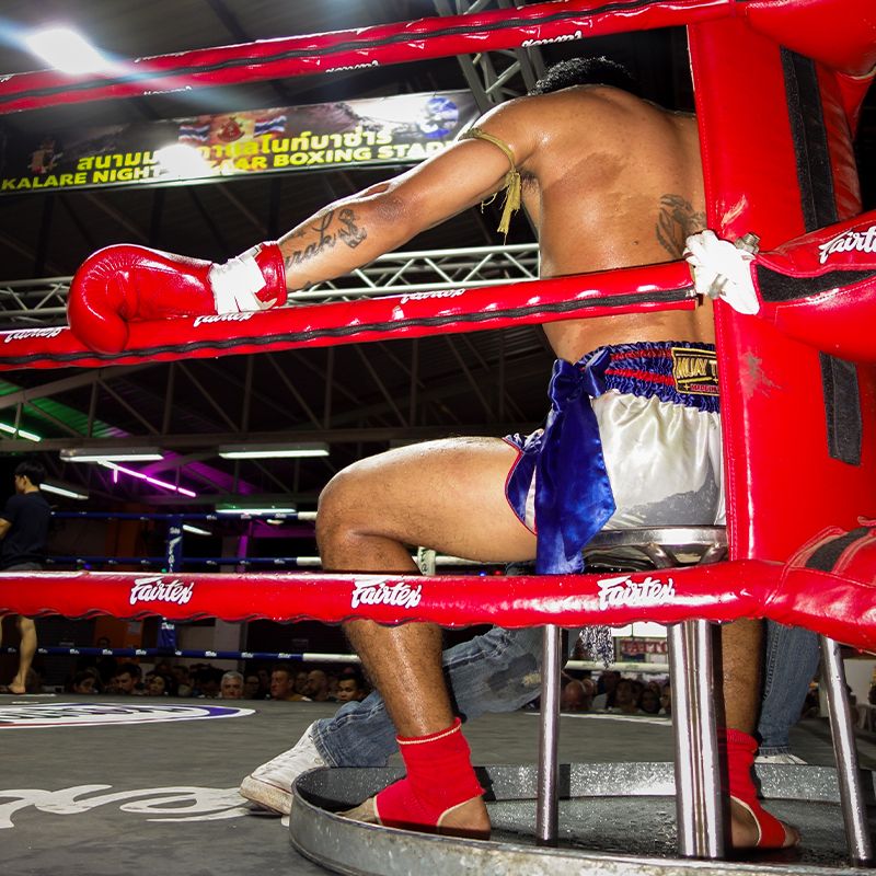 A Muay Thai fighter with red gloves rests on a stool between rounds in a boxing ring, under bright stadium lights.