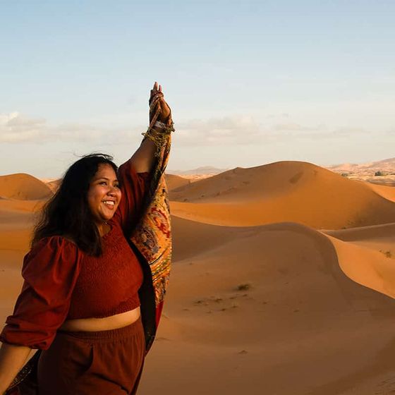 A woman joyfully holds up a patterned shawl, standing in a desert with orange sand dunes under a clear blue sky.