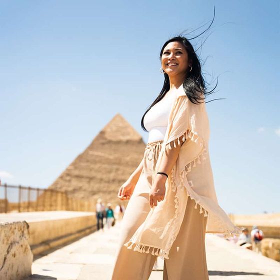 Woman smiling in front of the pyramids in Egypt, wearing a light beige outfit and standing on a stone pathway under a clear blue sky.