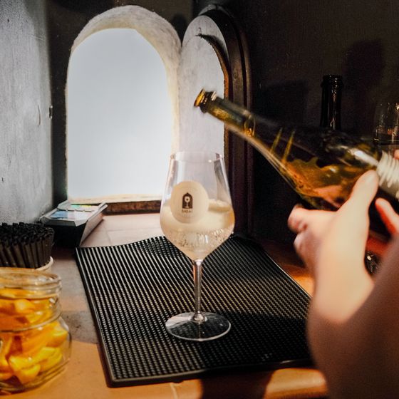 A person pours champagne into a branded wine glass on a black mat near a small window. A jar of lemons is nearby.