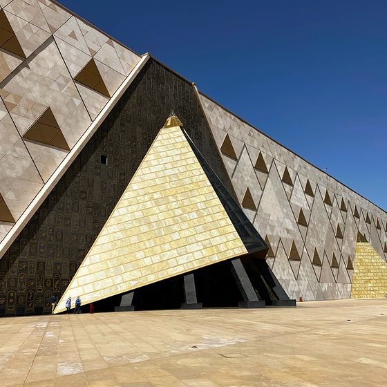 Modern architectural building with a large, golden triangular entrance and geometric facade patterns under a clear blue sky.