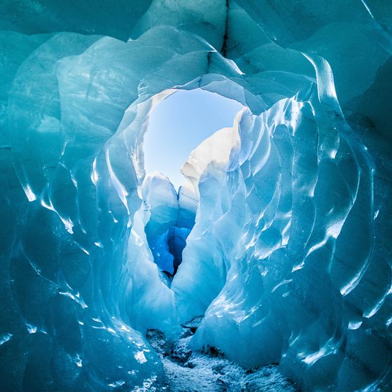 View through a glistening blue ice cave opening, revealing a bright sky beyond. The cave walls are textured with smooth, wavy patterns.