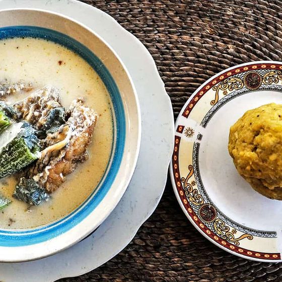A bowl of creamy soup with vegetables and meat next to a plate of fufu on a woven mat.