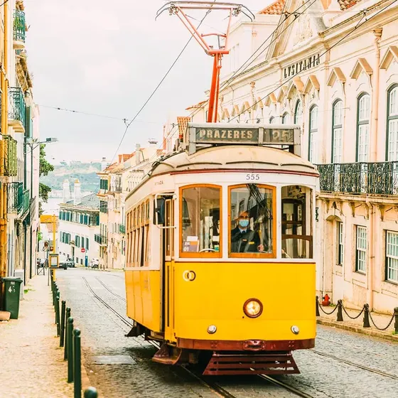 A bright yellow tram on a cobblestone street in Lisbon, with historic buildings and a view of a distant hill in the background.